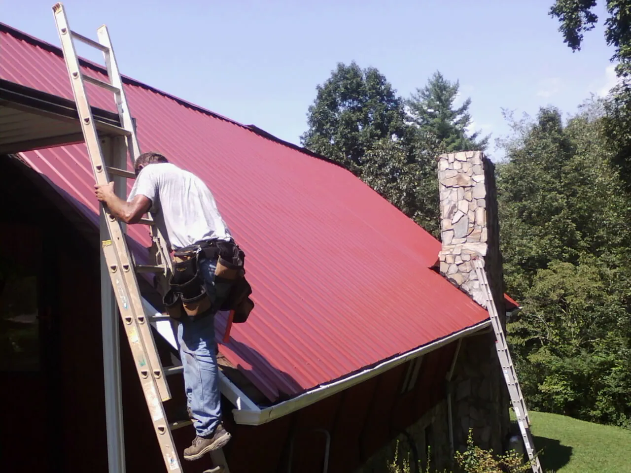 Roofer on a ladder beside a red metal roof and stone chimney — Soffit & Fascia Repair by Roofing Brothers in Eubank